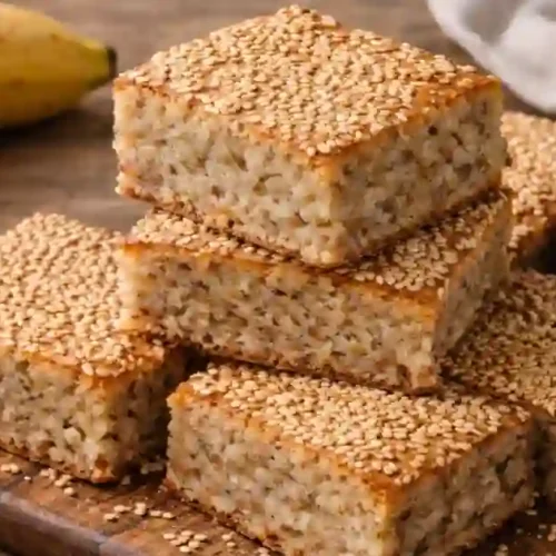 A stack of healthy sesame cake squares made without flour or sugar, garnished with sesame seeds, alongside bananas and a bowl of sesame seeds.