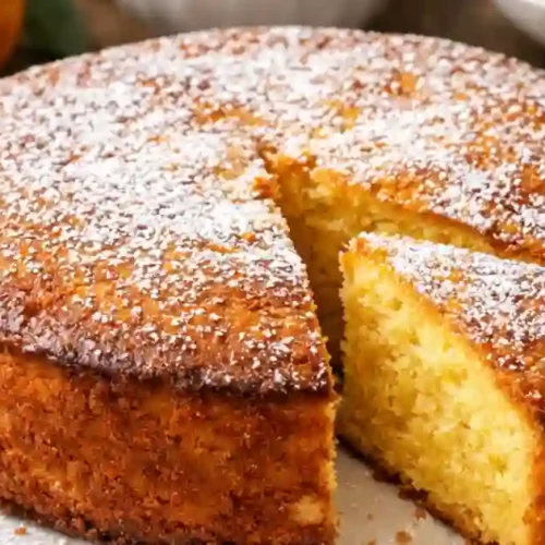 A freshly baked whole orange cake with a slice cut out, showcasing its moist texture and light color, dusted with powdered sugar.