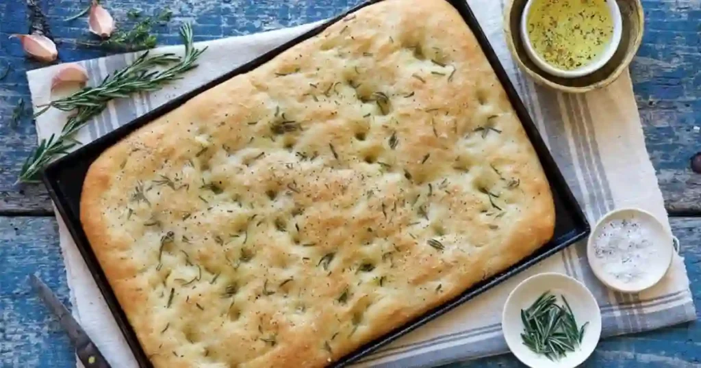 A beautifully risen overnight focaccia bread, golden brown and topped with fresh herbs, resting on a wooden board with a drizzle of olive oil nearby.