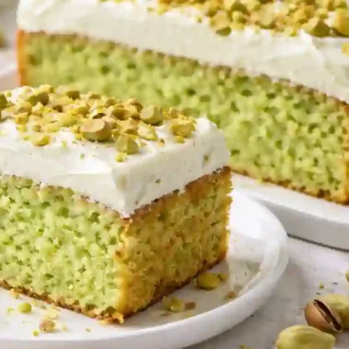 Slice of bright green pistachio pudding cake topped with white cream frosting and crushed pistachios, served on a white plate with scattered pistachios around.