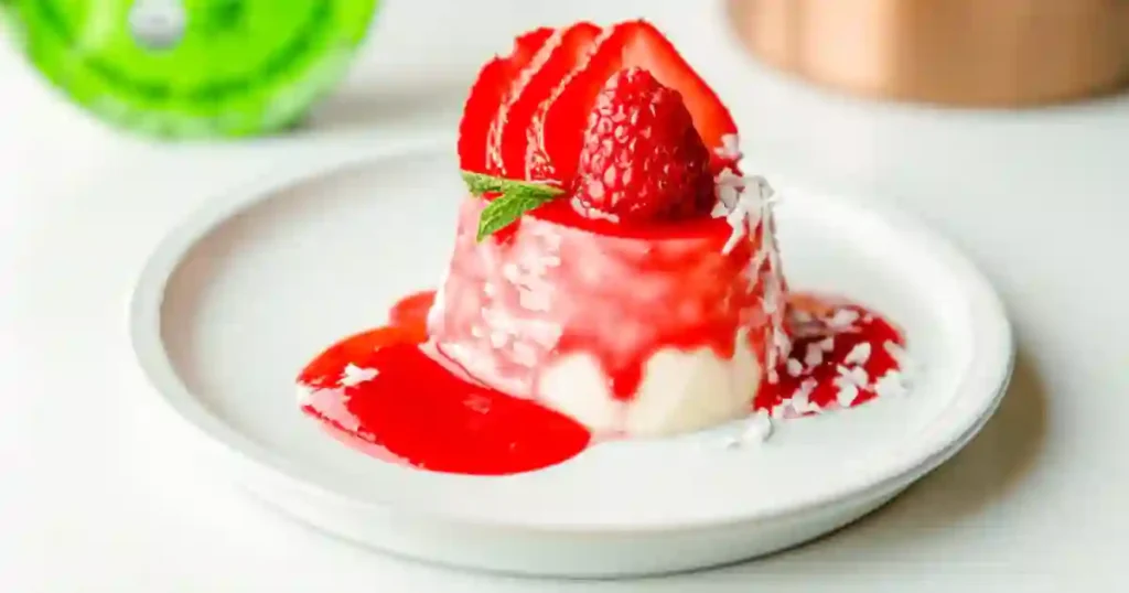 Delicious coconut milk panna cotta topped with fresh strawberries and raspberries, drizzled with berry sauce and garnished with shredded coconut and mint leaves.