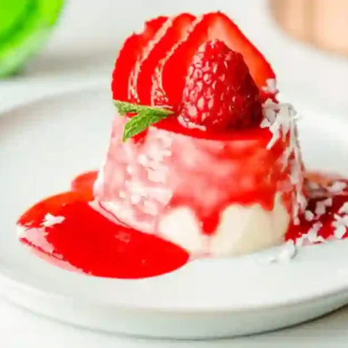 Delicious coconut milk panna cotta topped with fresh strawberries and raspberries, drizzled with berry sauce and garnished with shredded coconut and mint leaves.